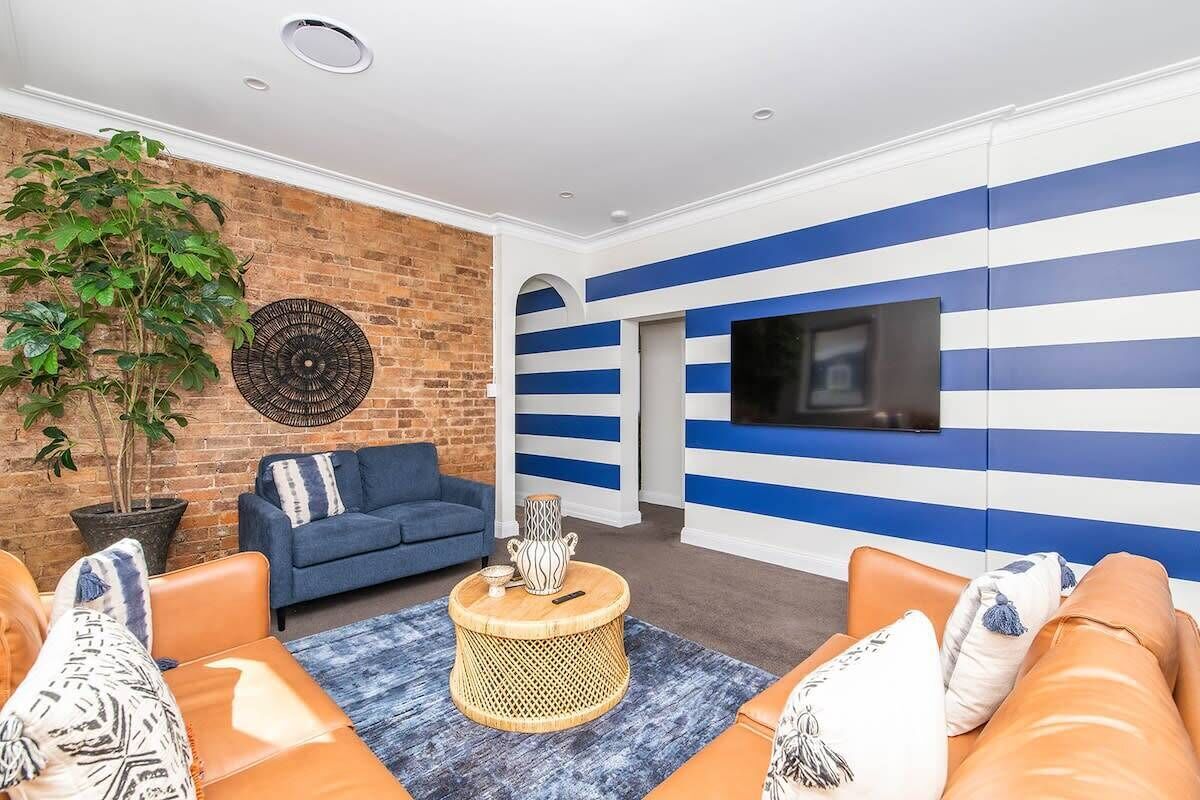 Ein Wohnzimmer mit Backsteinwand, blau-weiß gestreiften Wänden, einem Fernseher, Ledersofas und einer großen Pflanze.