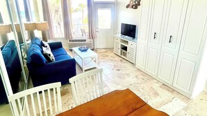 Una sala de estar con un sofá azul, una mesa de madera y un televisor. Se observan armarios blancos y un balcón con cortinas.