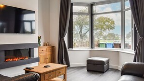 A modern living room with a fireplace, leather sofas, a wooden coffee table, and a large window with curtains. - 3 bedroom accommodation in St Annes (St Annes)