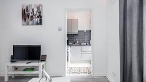 Una sala moderna con un televisor de pantalla plana sobre un mueble blanco, una mesita blanca y una escultura en espiral. Se ve una cocina a través de un acceso abierto, con armarios blancos y un salpicadero de azulejos.