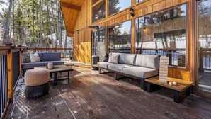 Eine Holzterrasse mit einer Couch und Hocker, großen Fenstern und Blick auf Bäume.