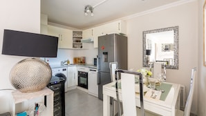 A modern kitchen with white cabinets, a stainless steel refrigerator, a dining table set with glassware, and a flat-screen TV mounted on the wall.