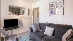 A living room with a gray sofa, a flat-screen TV, a glass coffee table, and a mirror with geometric patterns. There are four framed artworks on the wall.