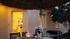 Un espace extérieur aménagé pour prendre les repas, avec une table dressée pour deux personnes, un panier de pain, une bouteille de vin et un vase de fleurs.
