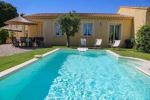 Piscina com espreguiçadeiras e uma mesa, rodeada por uma casa com telhado de azulejos e uma pequena árvore.