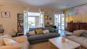 Amplia sala de estar con un sofá gris, sillones color beige y una mesa de centro blanca. Se observa una estantería y un ventanal con vistas.