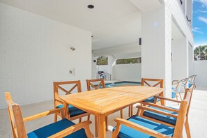 Ein Holztisch mit Stühlen und blauen Kissen in einem hellen, weiß verputzten Außenbereich.
