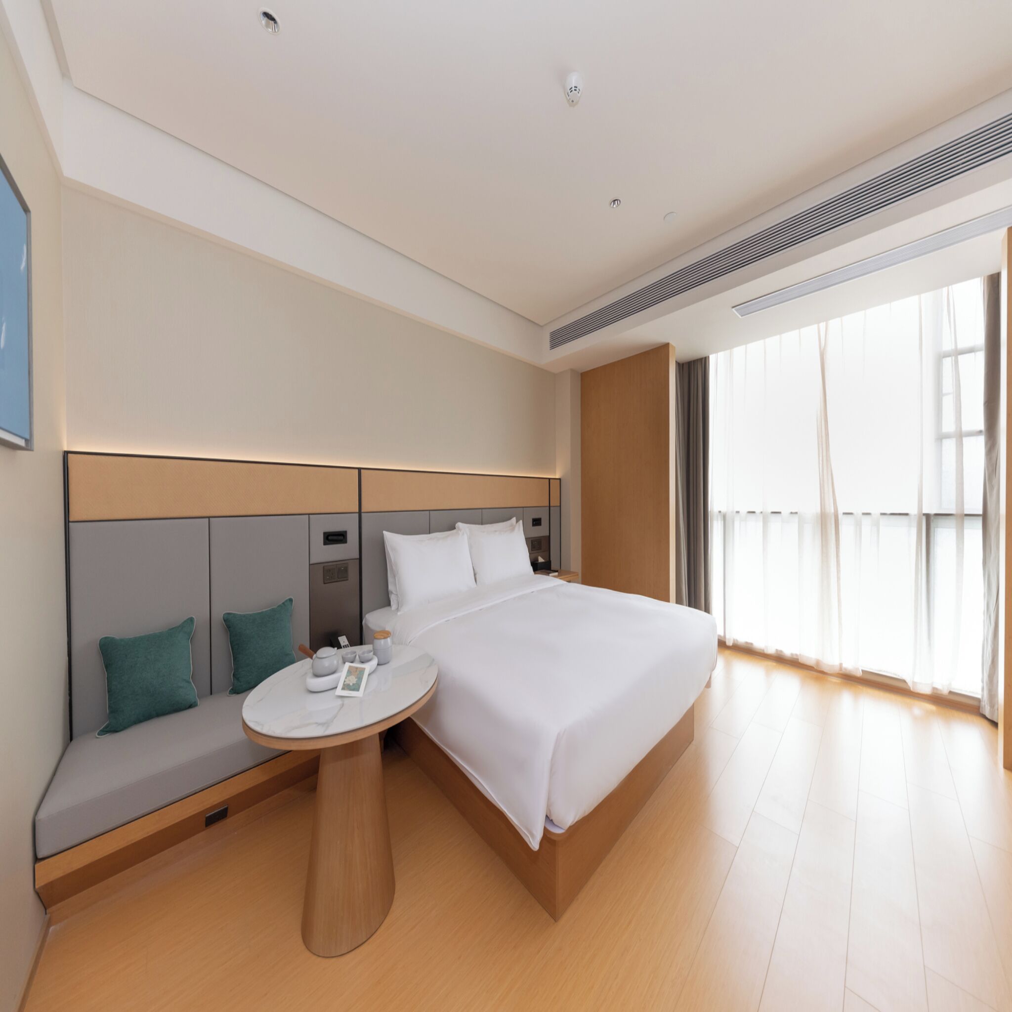 A modern hotel room with a large bed, a bedside table, a built-in bench with cushions, and a large window with curtains.