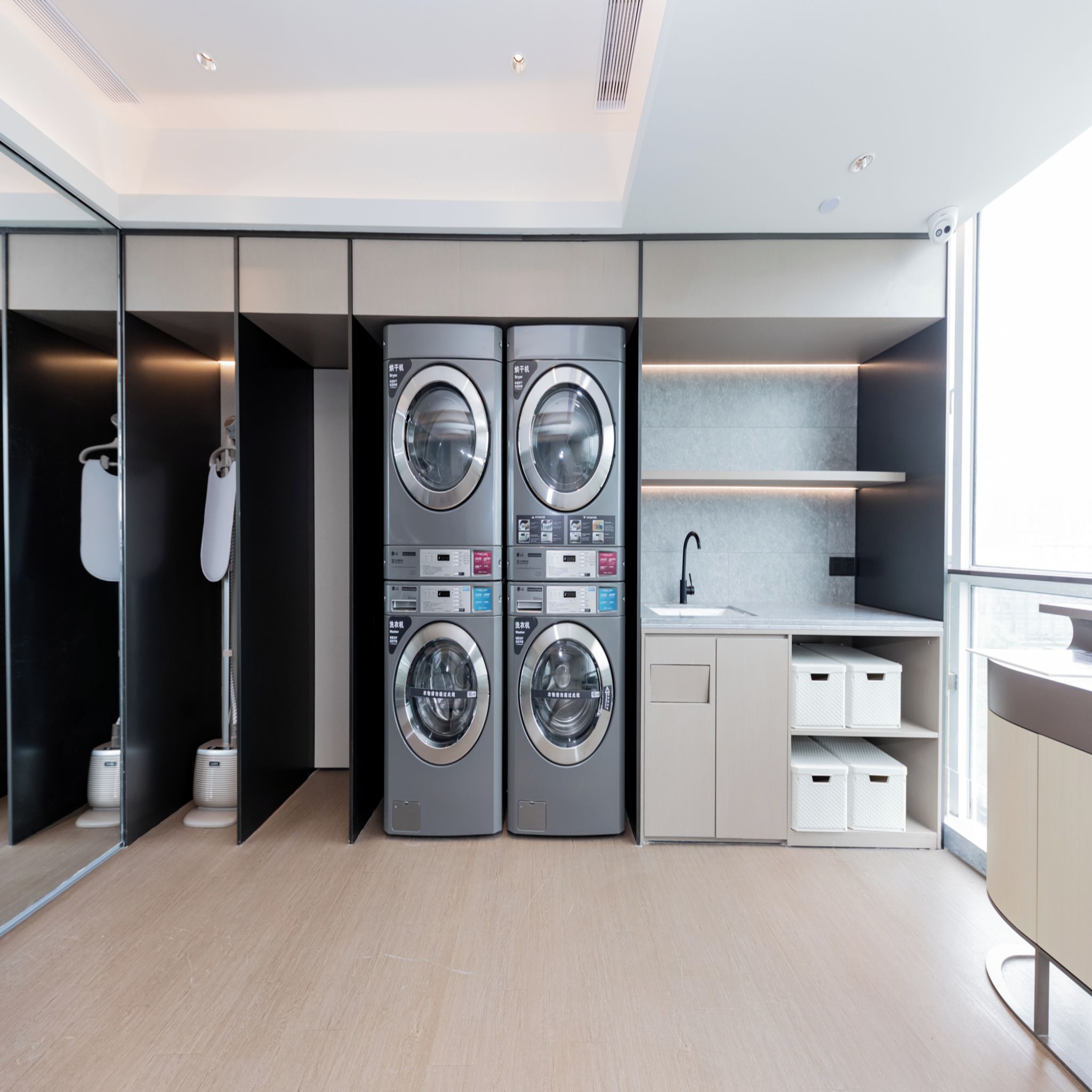 A modern laundry room with two stacked front-loading washing machines, a drying machine, a sink, and storage cabinets.