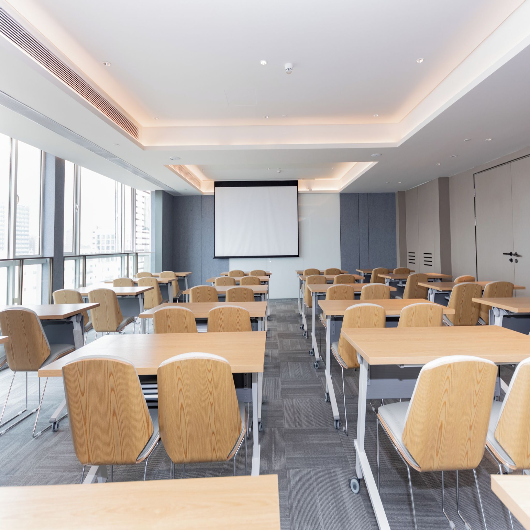 A modern classroom with rows of desks and chairs, a projector screen, and large windows.
