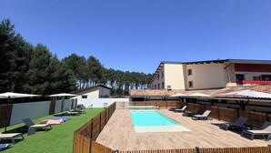 Zona de piscina con sillones de descanso, una terraza de madera y un edificio con techo de tejas.