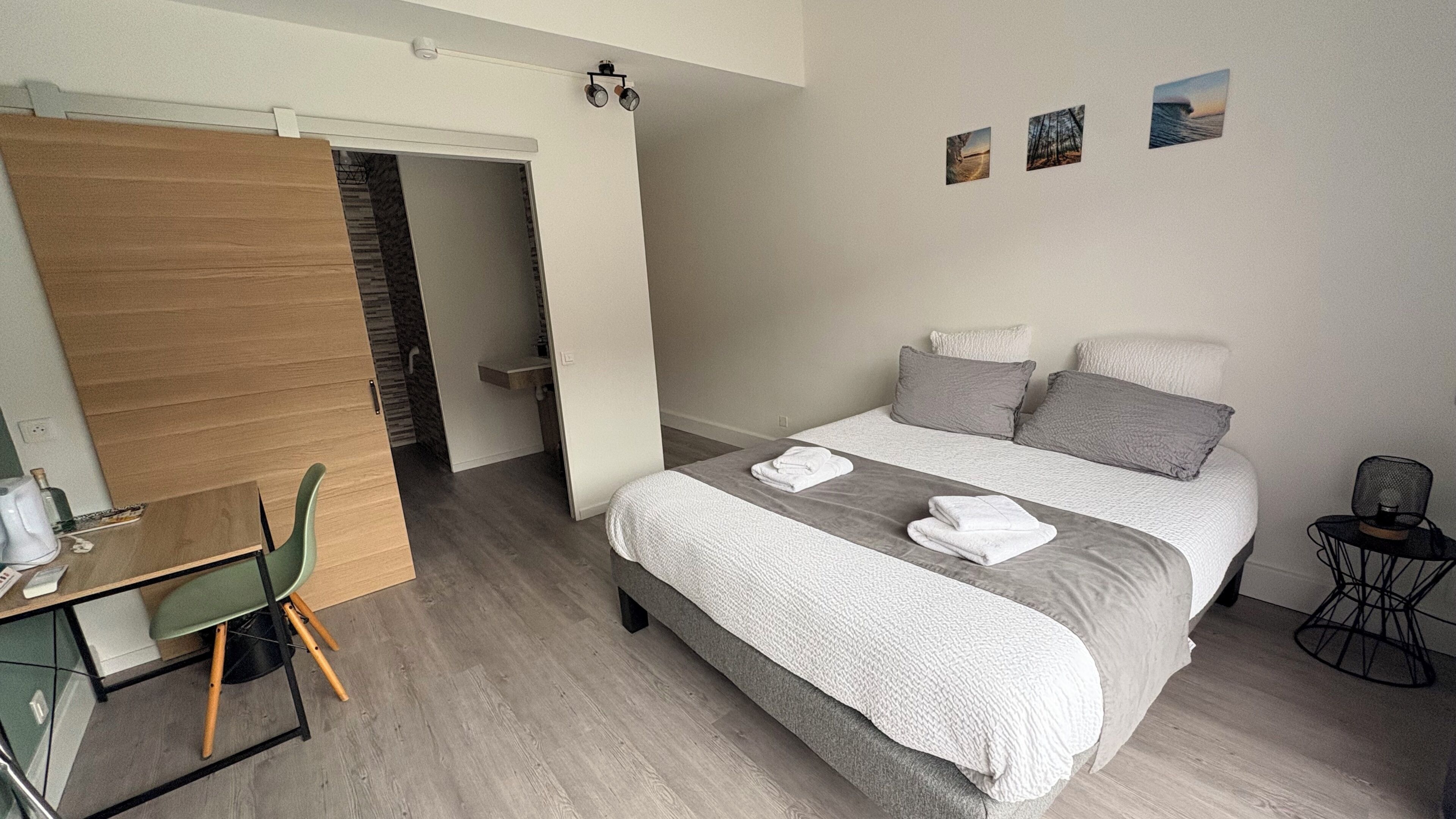 A hotel room with a bed, a desk, a chair, a small table, and a wall-mounted shelf.