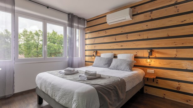 Un dormitorio moderno con una cama grande, paneles de madera en las paredes, una ventana con cortinas y un equipo de aire acondicionado.
