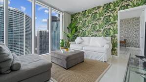 Een moderne woonkamer met een grijze bank, een grijze poef en een witte divan. Er is een groot raam met uitzicht op hoge gebouwen. De kamer heeft tropisch behang en een kleine plant op de poef.