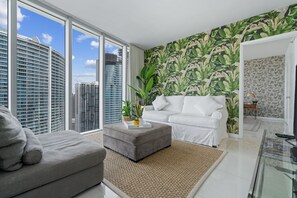 Una sala de estar moderna con un sofá gris, un reposapiés gris y un sofá blanco. Hay un ventanal con vistas a edificios altos. La habitación tiene un papel tapiz tropical y una planta pequeña sobre el reposapiés.