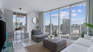 Een moderne woonkamer met een groot raam, een grijze bank, een poef en een flatscreen televisie.