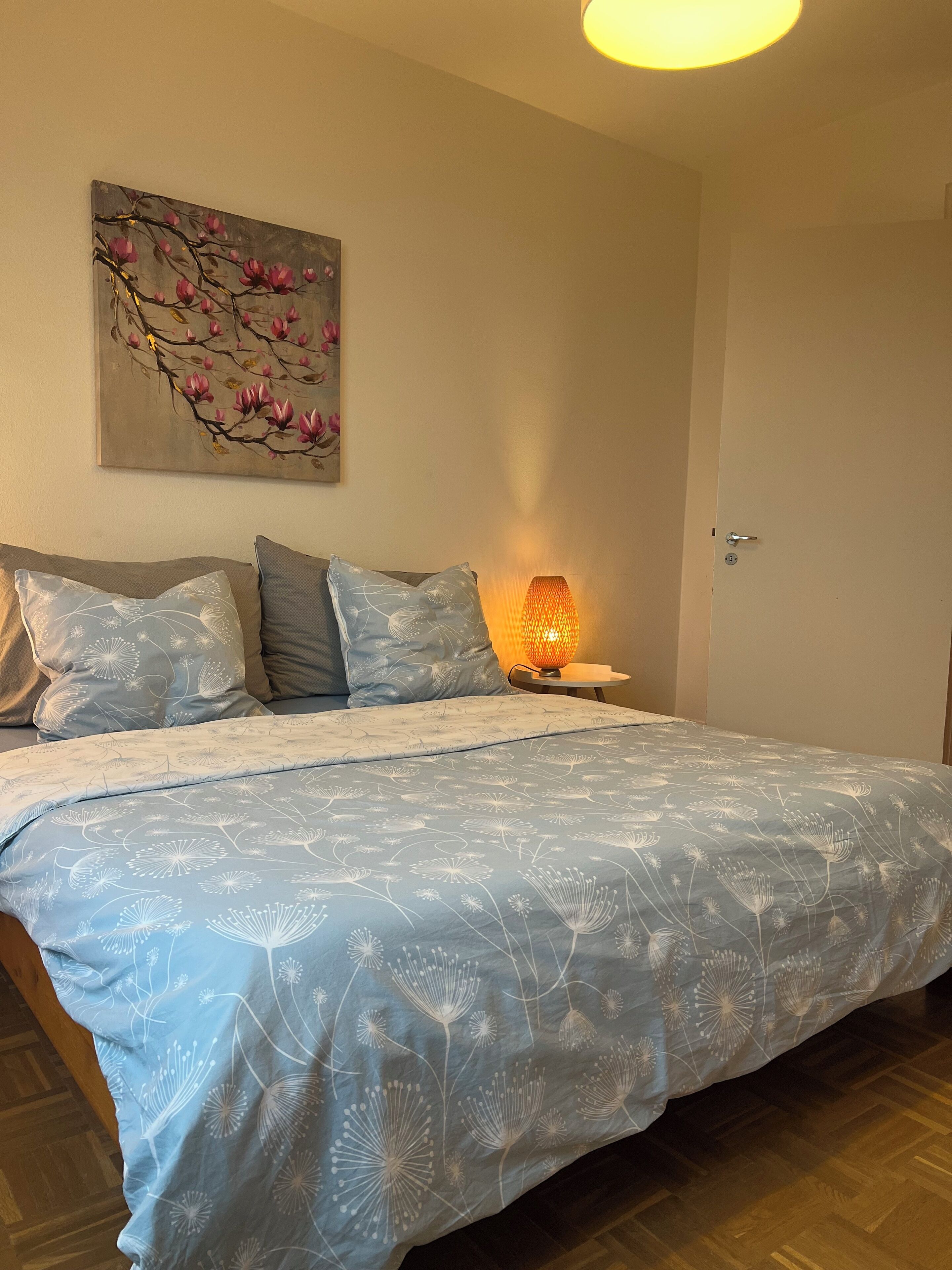 Een slaapkamer met een bed, kussens, een nachtkastje, een lamp en een schilderij aan de muur.