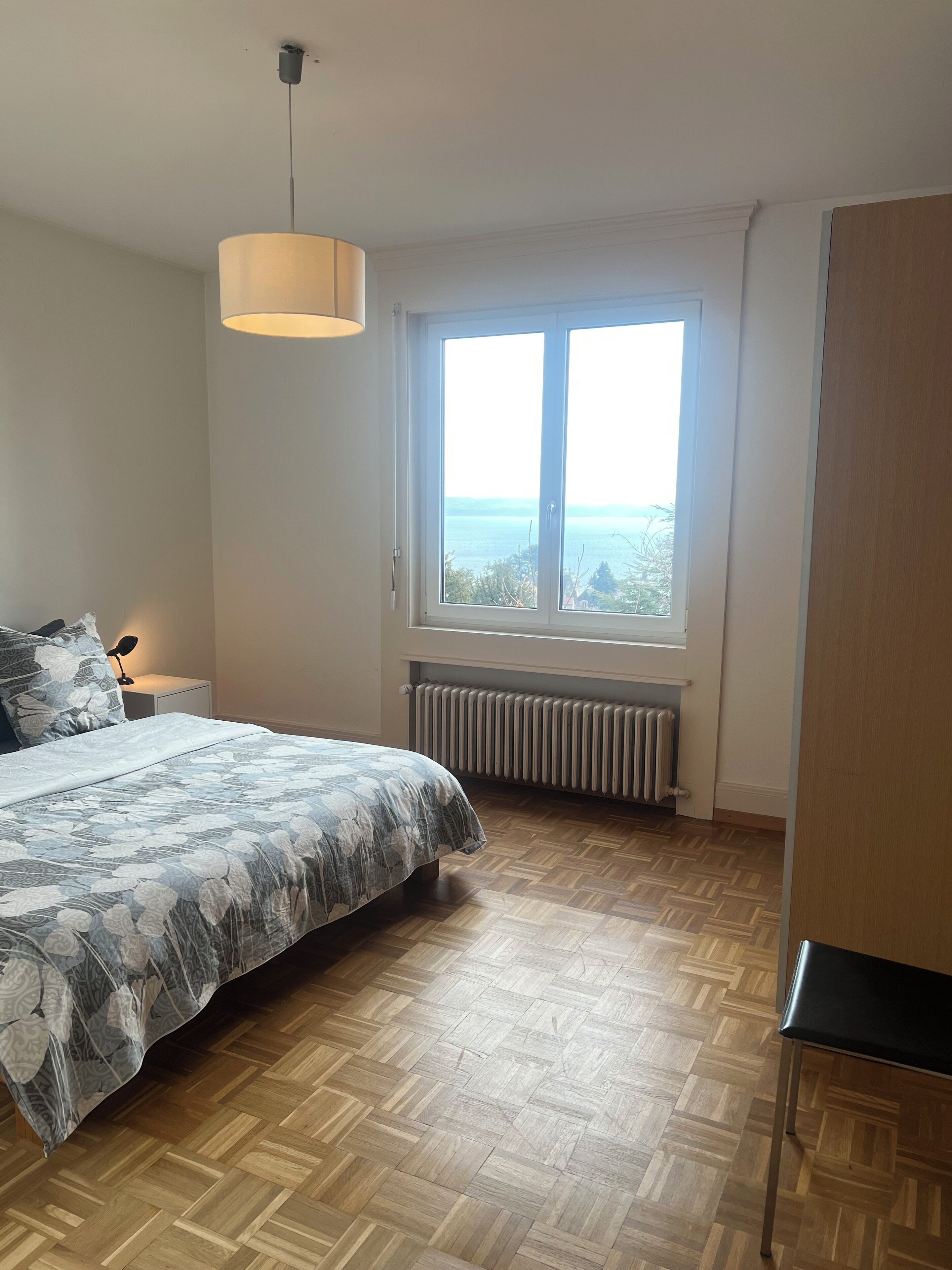 Ein Schlafzimmer mit einem Bett, einem Fenster mit Blick, einem Heizkörper und einer Pendelleuchte.