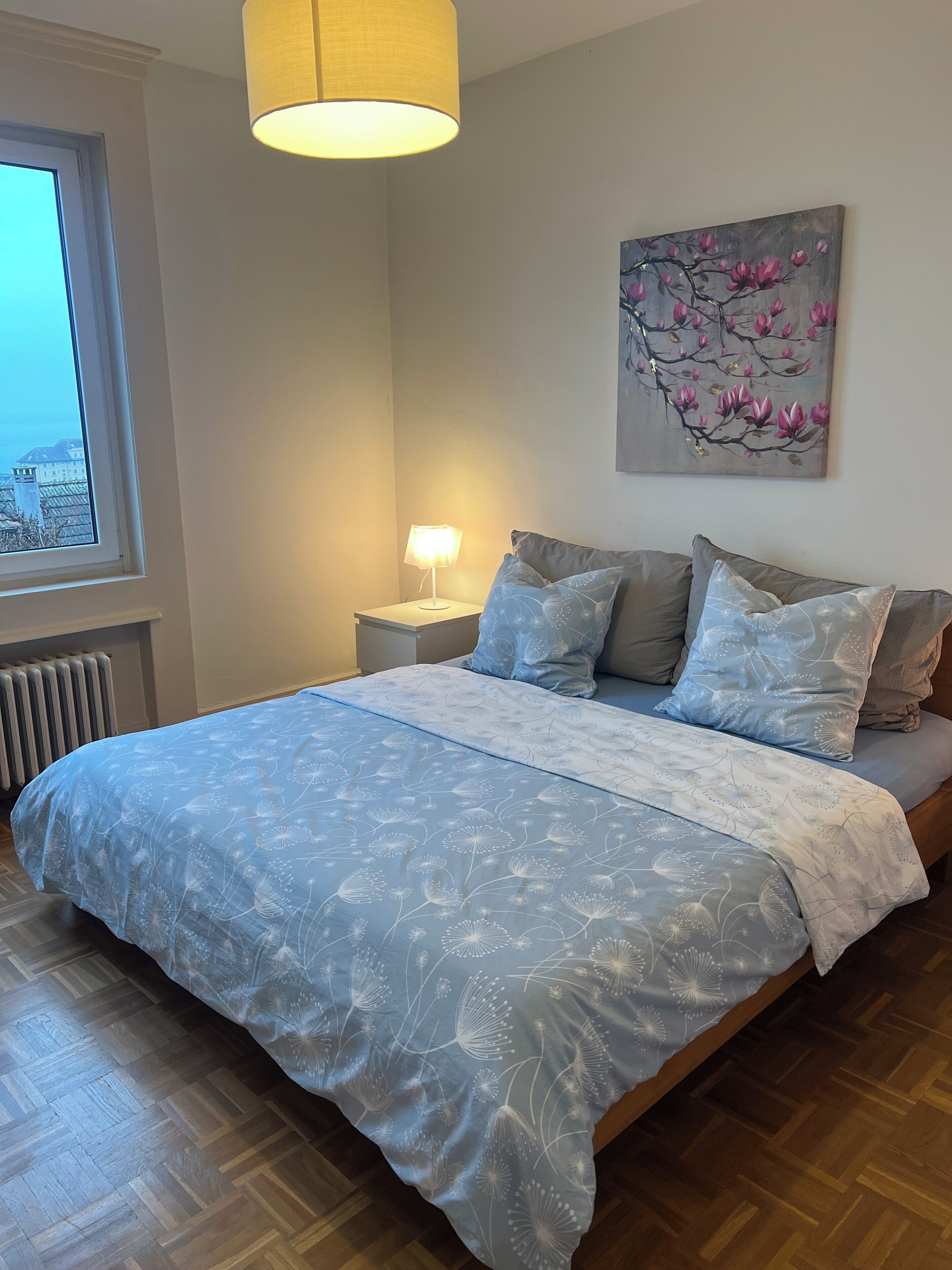 Une chambre à coucher avec un lit, une table de chevet, une lampe, une fenêtre et un tableau au mur.