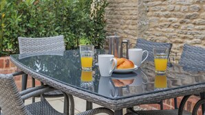 Una mesa de vidrio con alimentos para el desayuno, como croissants, café y jugo de naranja, frente a una pared de piedra y follaje.