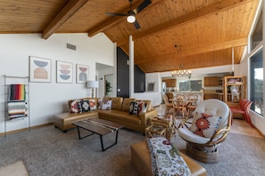 Un salón espacioso con techo alto, vigas de madera, un sofá de cuero marrón, un sillón estampado y un comedor con muebles de madera.