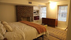 A bedroom with a brick fireplace, a bed with white bedding, a wooden dresser, and a patterned rug. - Elegant Savannah Patio Apartment! An Arty Flair on Jones Street (Savannah)