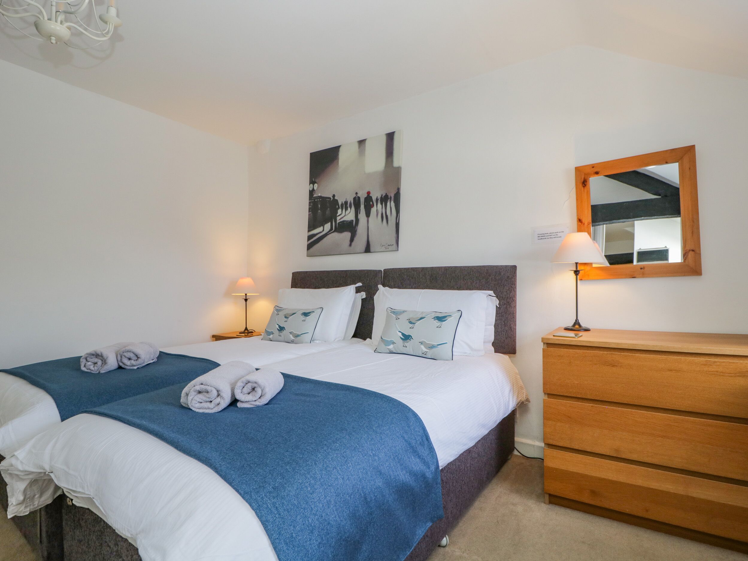 A double bed with two pillows, a bedside table with a lamp, a wooden dresser, and a wall-mounted mirror.