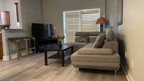 A modern living room with a sectional sofa, a coffee table, a TV, and a kitchen area with a countertop and bar stools. - Paradise Villa in Regal Palms (Davenport)