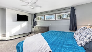 A bedroom with a bed, a ceiling fan, a dresser, a TV mounted on the wall, and a window with curtains.