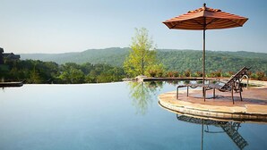 A swimming pool with a lounge chair and umbrella, surrounded by a landscaped area with a view of hills. - Family Fun at Wilderness Club at Big Cedar! (Ridgedale)