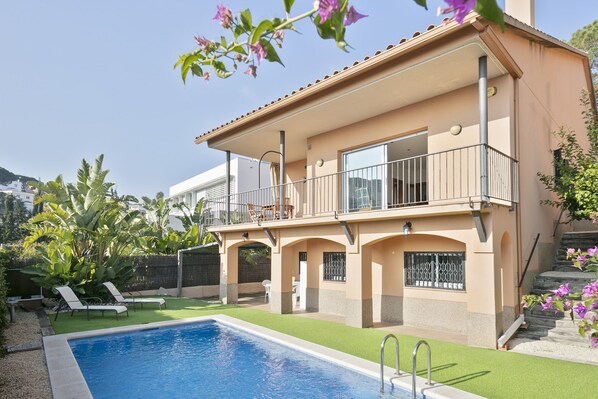 A two-story house with a swimming pool, a balcony, and a garden with tropical plants.