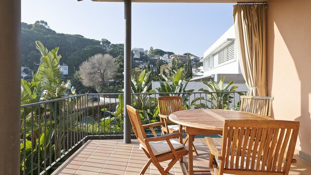 A balcony with a wooden table and chairs, surrounded by a metal railing, overlooking a lush green landscape with trees and buildings in the background.