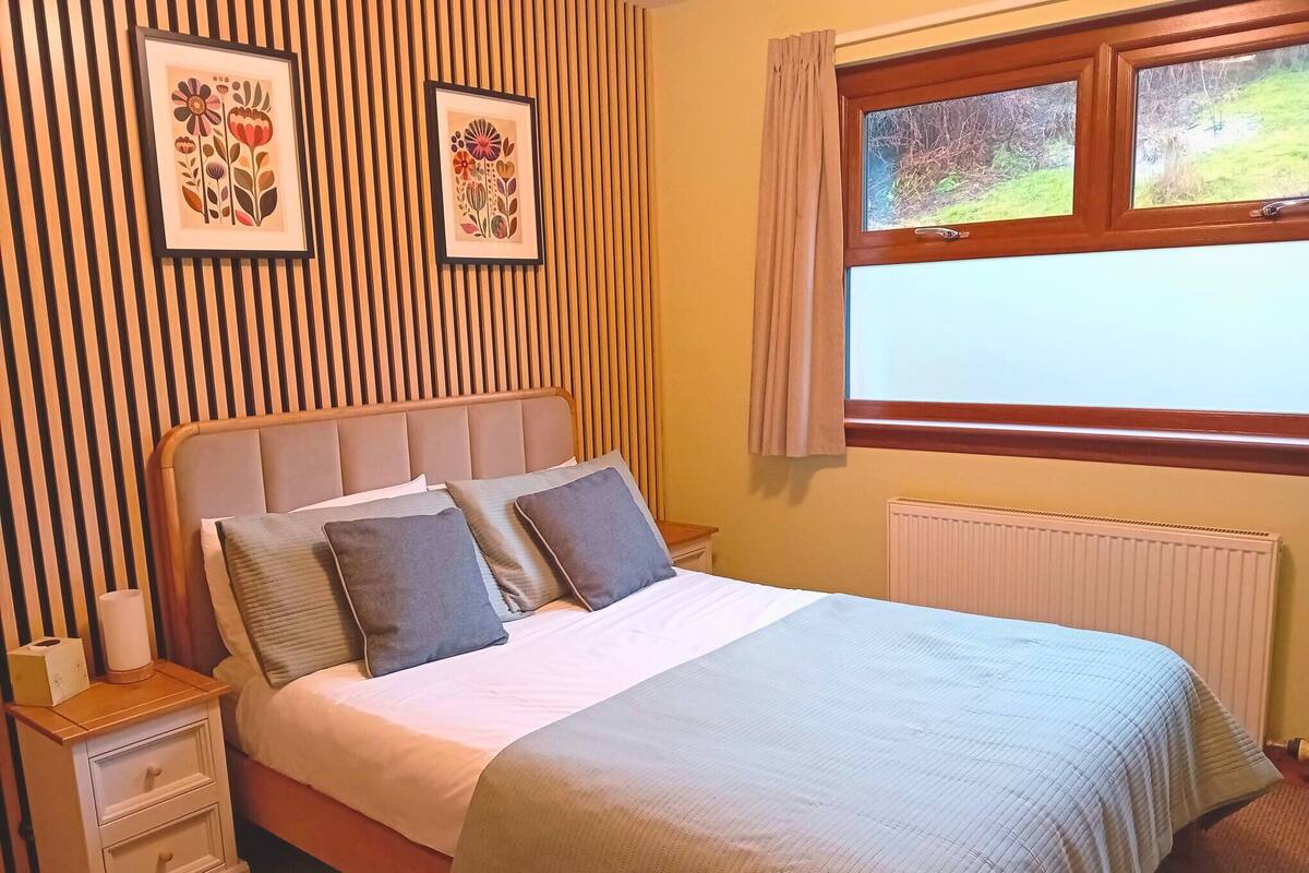 Un lit bien fait, avec une couette grise et blanche, une tête de lit en bois et deux tables de chevet avec des lampes. Deux tableaux encadrés représentant des motifs floraux ornent le mur, et une fenêtre est agrémentée de rideaux.