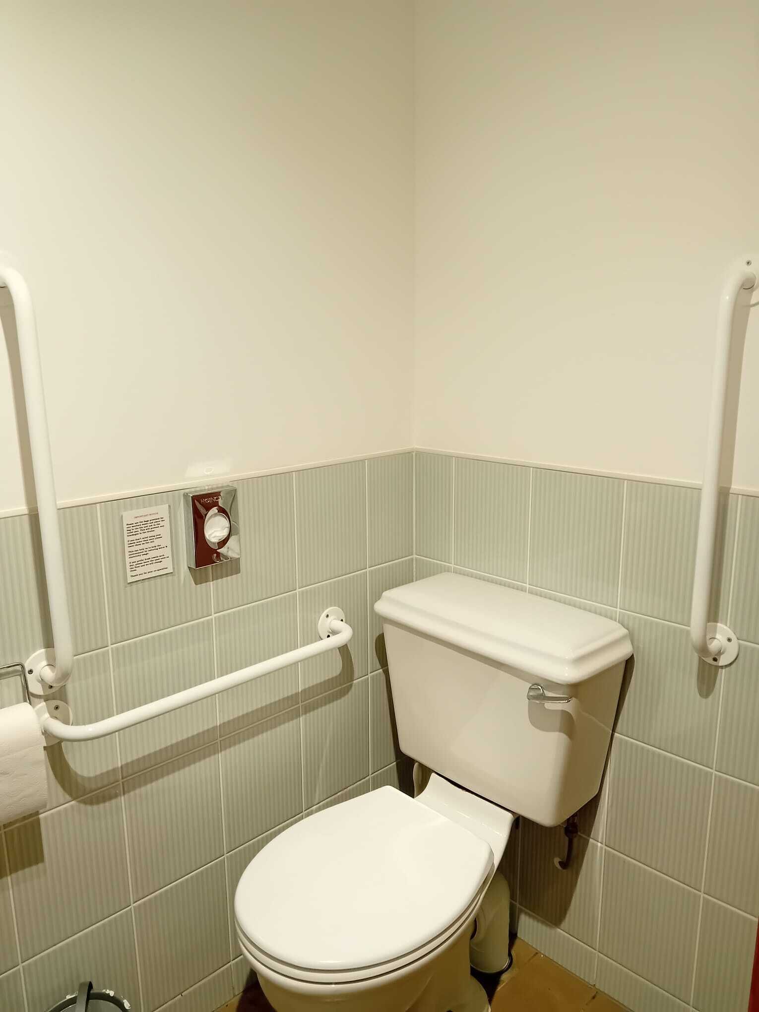 A small, single-occupancy bathroom with a white toilet, a handrail, and a toilet paper holder.