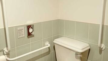 A small, single-occupancy bathroom with a white toilet, a handrail, and a toilet paper holder.