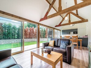 A spacious living area with a wooden ceiling, large windows, a sofa, a coffee table, and a kitchen in the background.