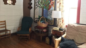 A cozy room with a wooden floor, a green armchair, a small table with a lamp, a wall tapestry, and a window with curtains. - "Little House on the Prairie" with modern plumbing! (Coon Valley)