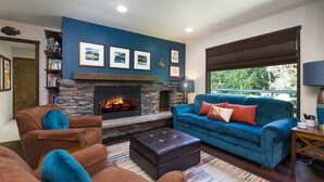 A living room with a stone fireplace, blue walls, a blue sofa, and a view of greenery outside. - Well-furnished Hiker's Retreat, Centrally located for visiting the ONP; Sleeps 8 (Port Angeles)