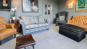 Ein Wohnzimmer mit grauem Sofa, einem braunen Ledersessel, einem grauen Sessel, einem Holztisch und einem schwarzen Hocker. An den Wänden befinden sich Bilder und eine Tür mit Glasverzierungen.