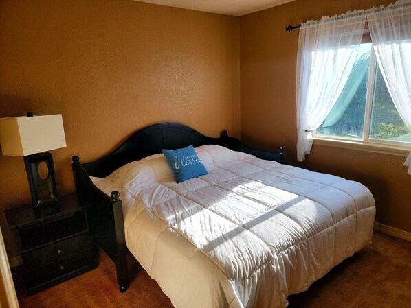 A neatly made bed with a white comforter and a blue pillow, a bedside table with a lamp, and a window with white curtains.