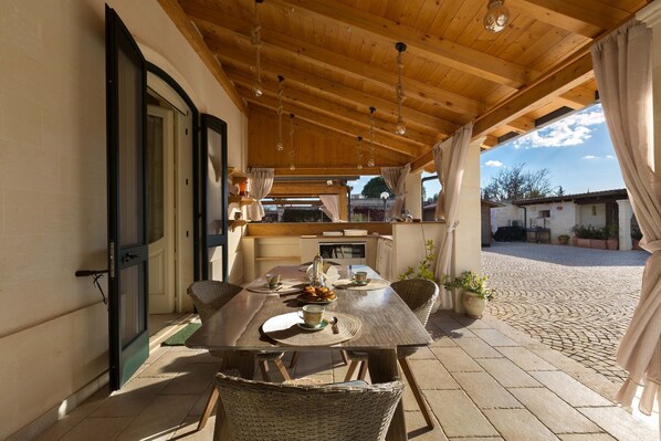 An outdoor dining area with a wooden table and chairs, a view of the courtyard, and a covered seating area with a wooden ceiling and hanging lights. - Villa Nicrys by Perle di Puglia (Maglie)