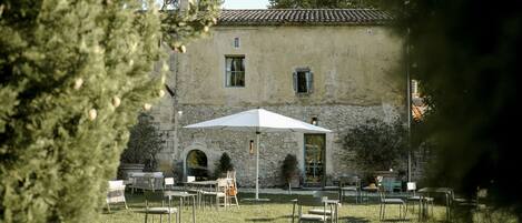 An outdoor seating area with tables and chairs, a stone building with a tiled roof, and a large umbrella providing shade.