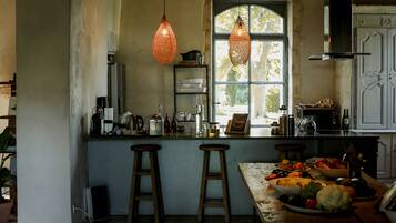 Une cuisine avec des tabourets en bois, une grande fenêtre, des suspensions décoratives et divers ustensiles de cuisine disposés sur le plan de travail.