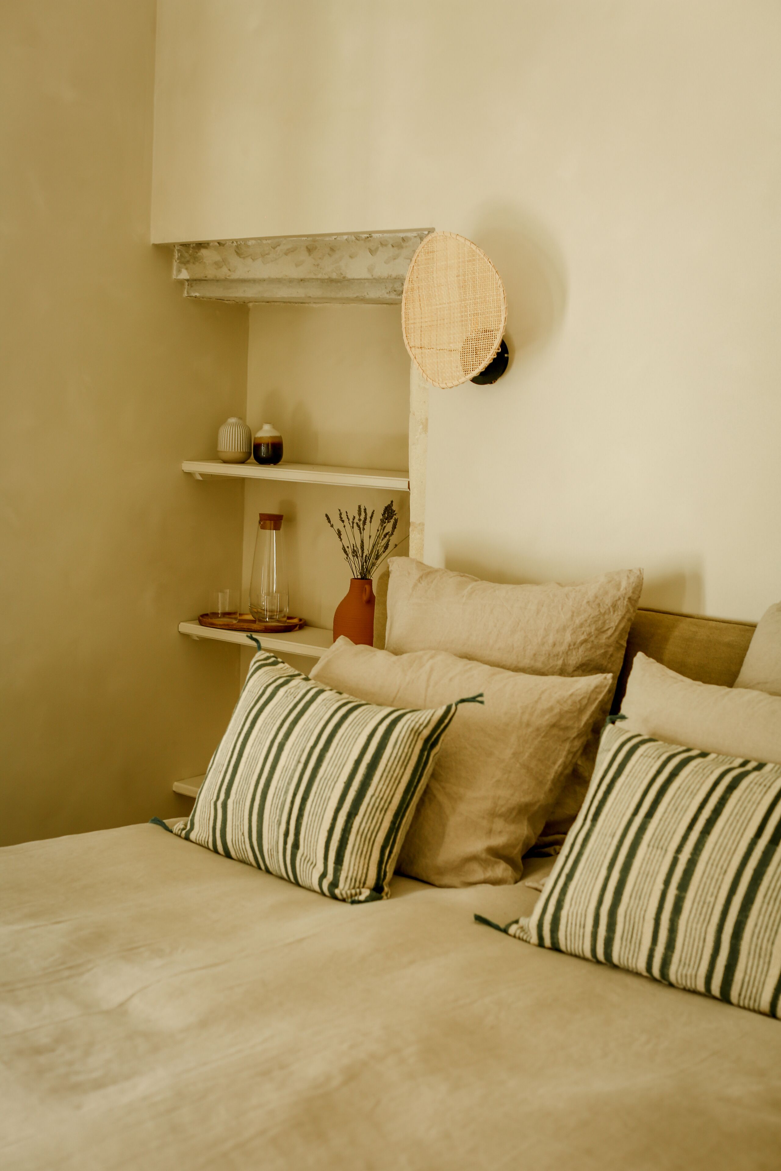 Un dormitorio con cama, almohadas, una mesita de noche con un florero y una estantería con objetos decorativos.