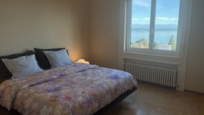 Une chambre à coucher avec un lit, une fenêtre donnant sur un lac et des bâtiments, et un radiateur placé sous la fenêtre.