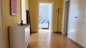 Un couloir doté d’un plancher en bois, d’une commode blanche, d’un miroir et d’une porte donnant sur une chambre à coucher avec un lit et une table de chevet.