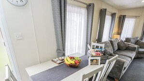 A dining area with a table set for two, a bowl of fruit, a tray of snacks, and a card with a message. - The Cotswold Holiday Lodge (Evesham)