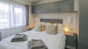 A bedroom with a bed, grey cabinets, a window with curtains, and a bedside lamp. - The Cotswold Holiday Lodge (Evesham)