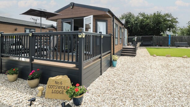 A lodge with a deck, a gravel path, and a sign indicating it's No. 6 Cotswold Lodge. - The Cotswold Holiday Lodge (Evesham)