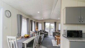 A compact living space with a dining area, a sofa set, a kitchen with a microwave, and a view to the outside. - The Cotswold Holiday Lodge (Evesham)